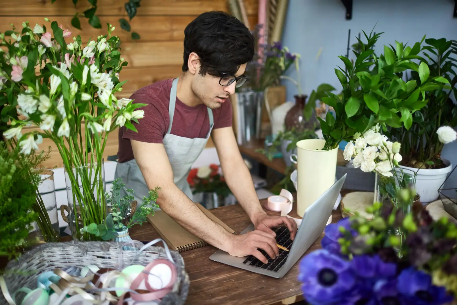 A florist in an apron types on a laptop at his shop's worktable, surrounded by vases of fresh flowers