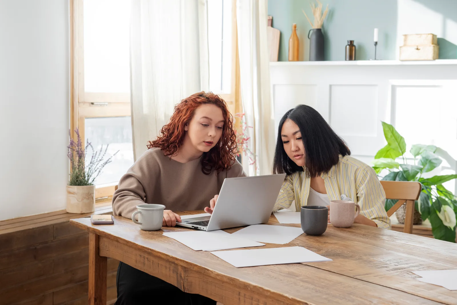 Two women compare notes on a laptop at a wooden table, papers and coffee mugs spread around them