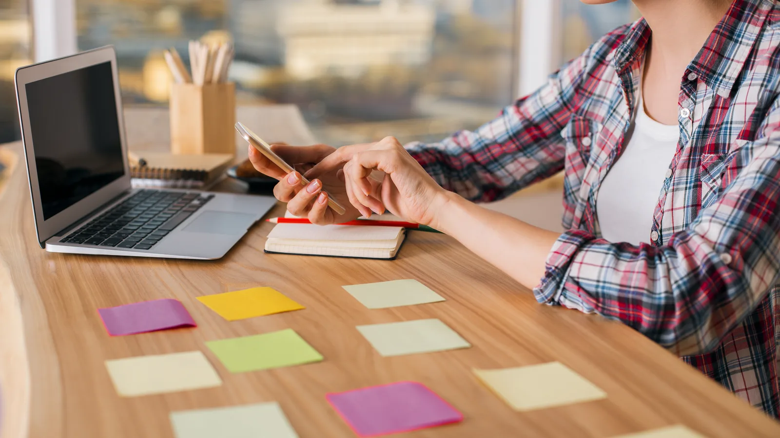 A woman checks her phone beside an open laptop, colored sticky notes laid out in rows across her desk