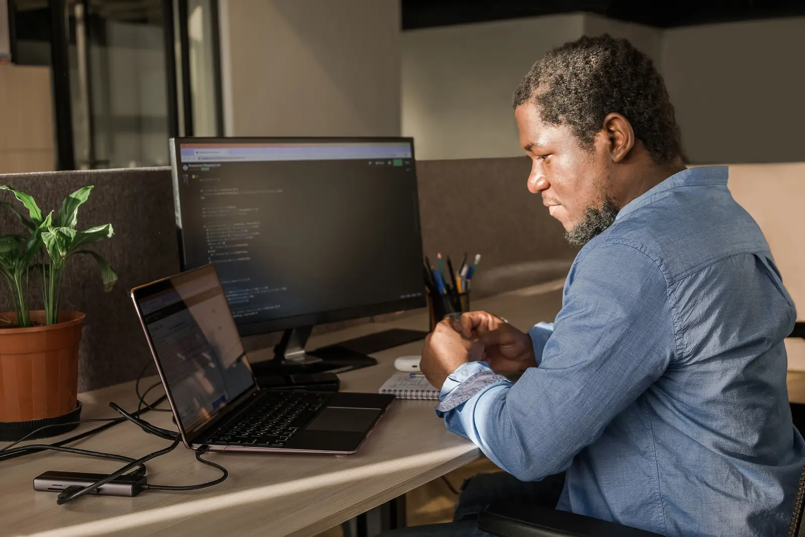 A developer in a denim shirt reads code on a desktop monitor, his laptop open beside it at an office desk