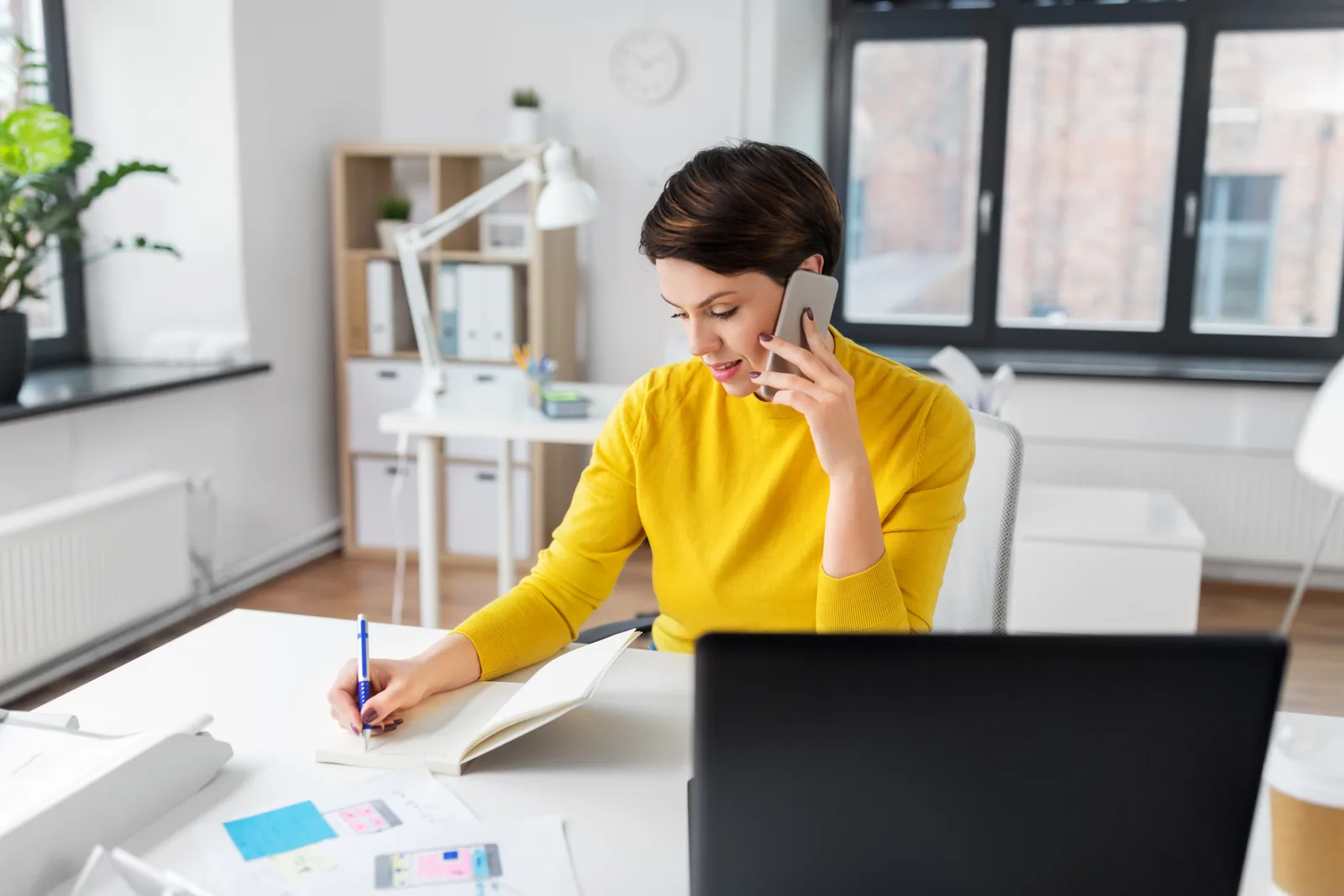 A woman on a call at her desk takes notes beside her laptop, the voice channel in practice