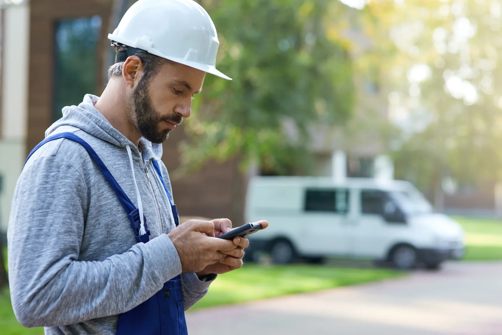 A builder in a hard hat and overalls reads his phone outside, a white work van parked behind him