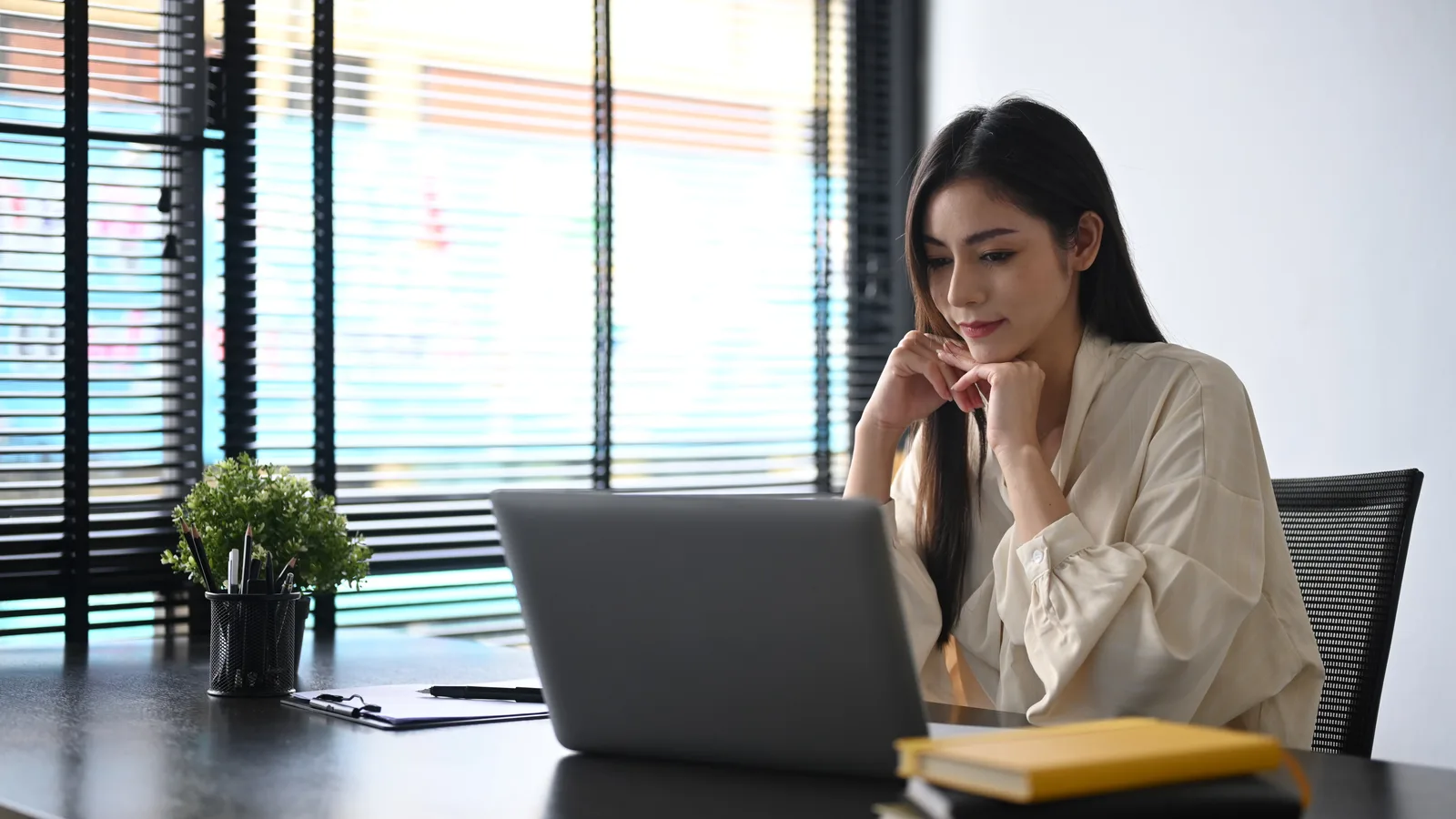 A small business owner at her office desk reads her laptop screen, chin resting on her hands