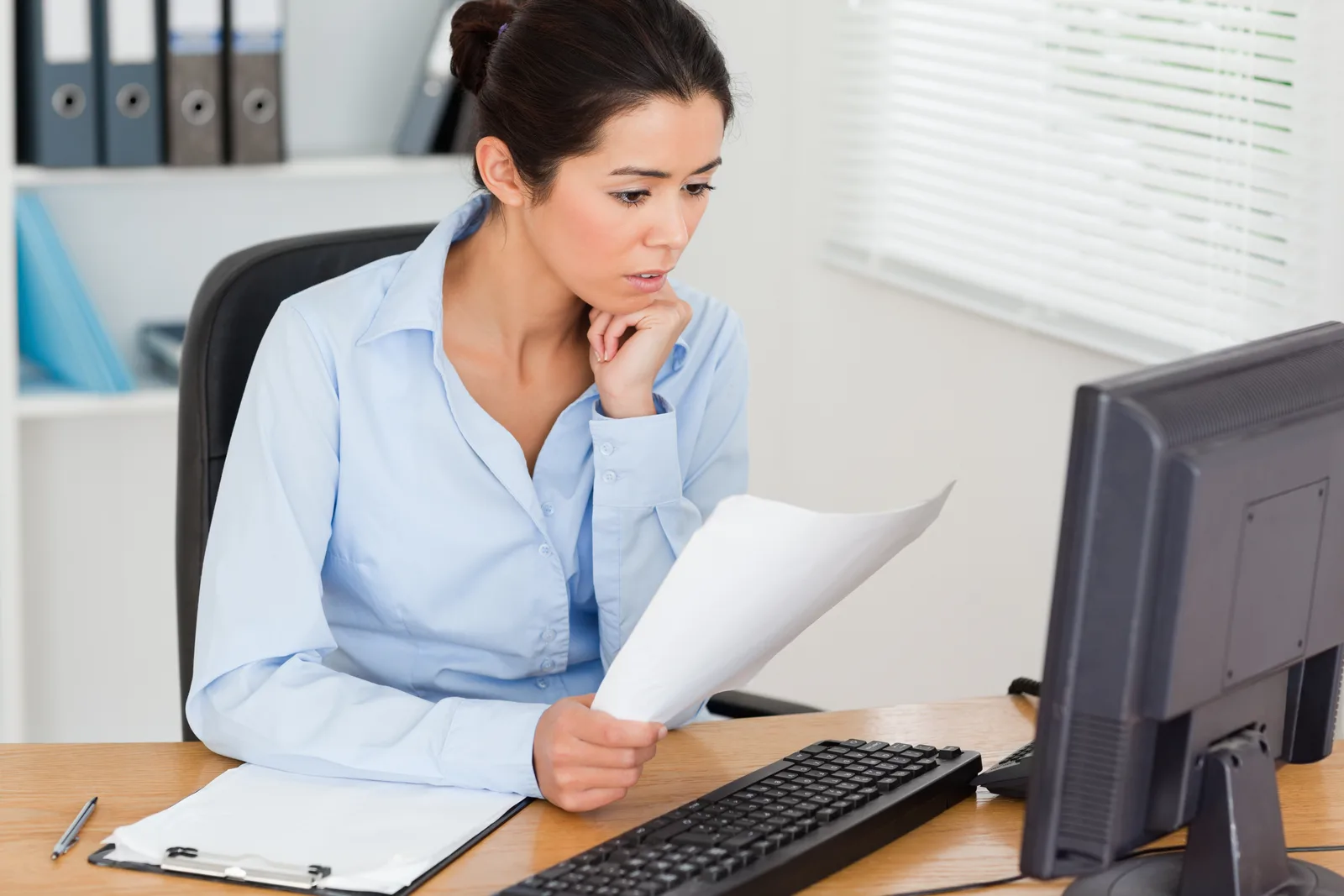An office manager holds a printed document and checks it against her computer screen