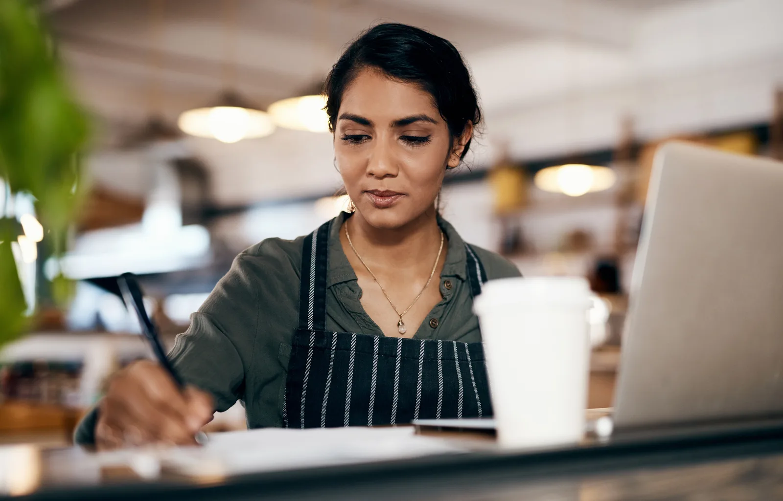 A café owner in an apron writes down figures beside her laptop and a coffee cup at the counter
