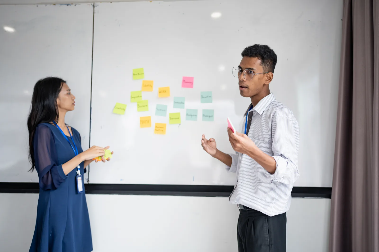 Two coworkers talk through a plan at an office whiteboard covered in sticky notes