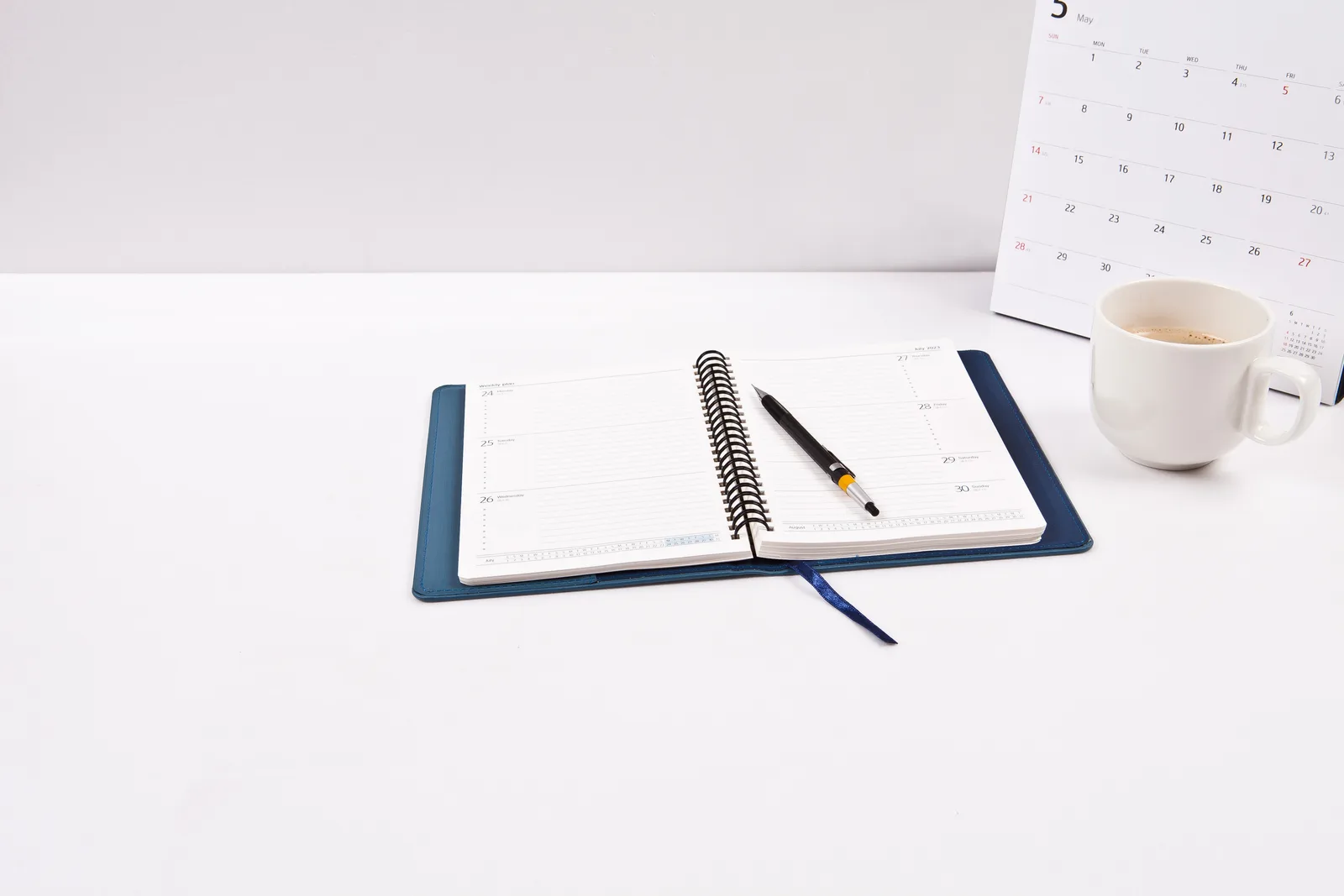 An open weekly planner and pen beside a desk calendar and a cup of coffee