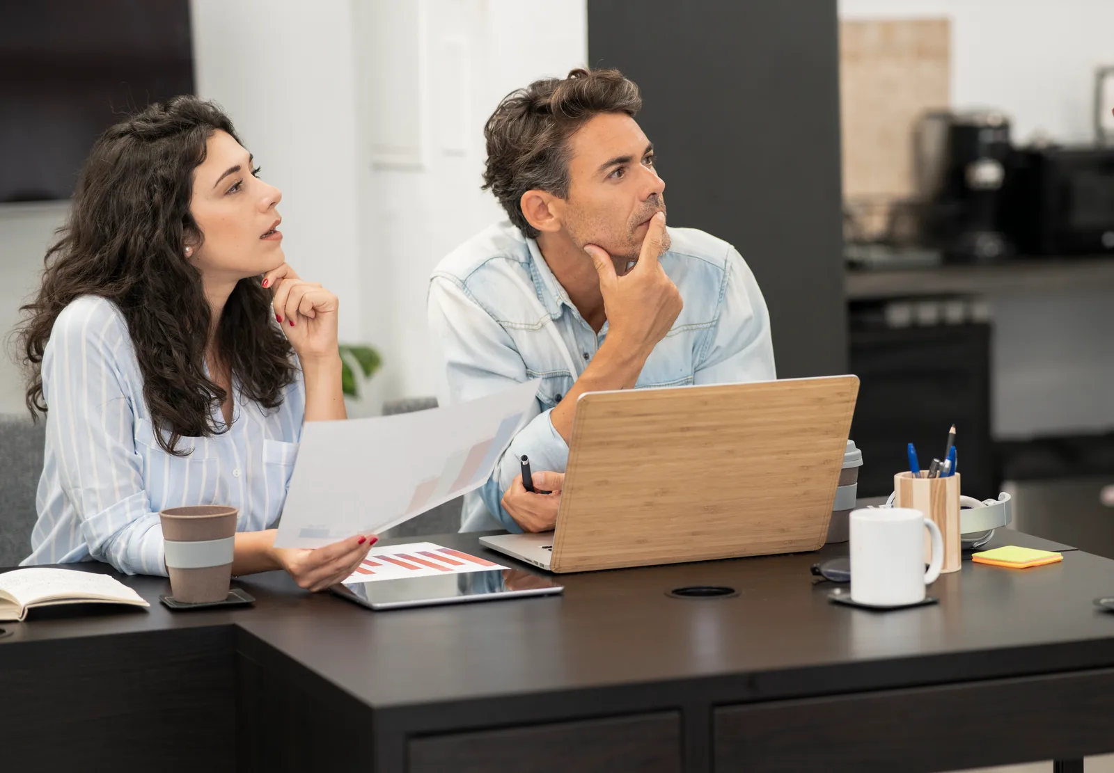 Two colleagues at a desk weighing a decision, one holding printed pages beside a laptop