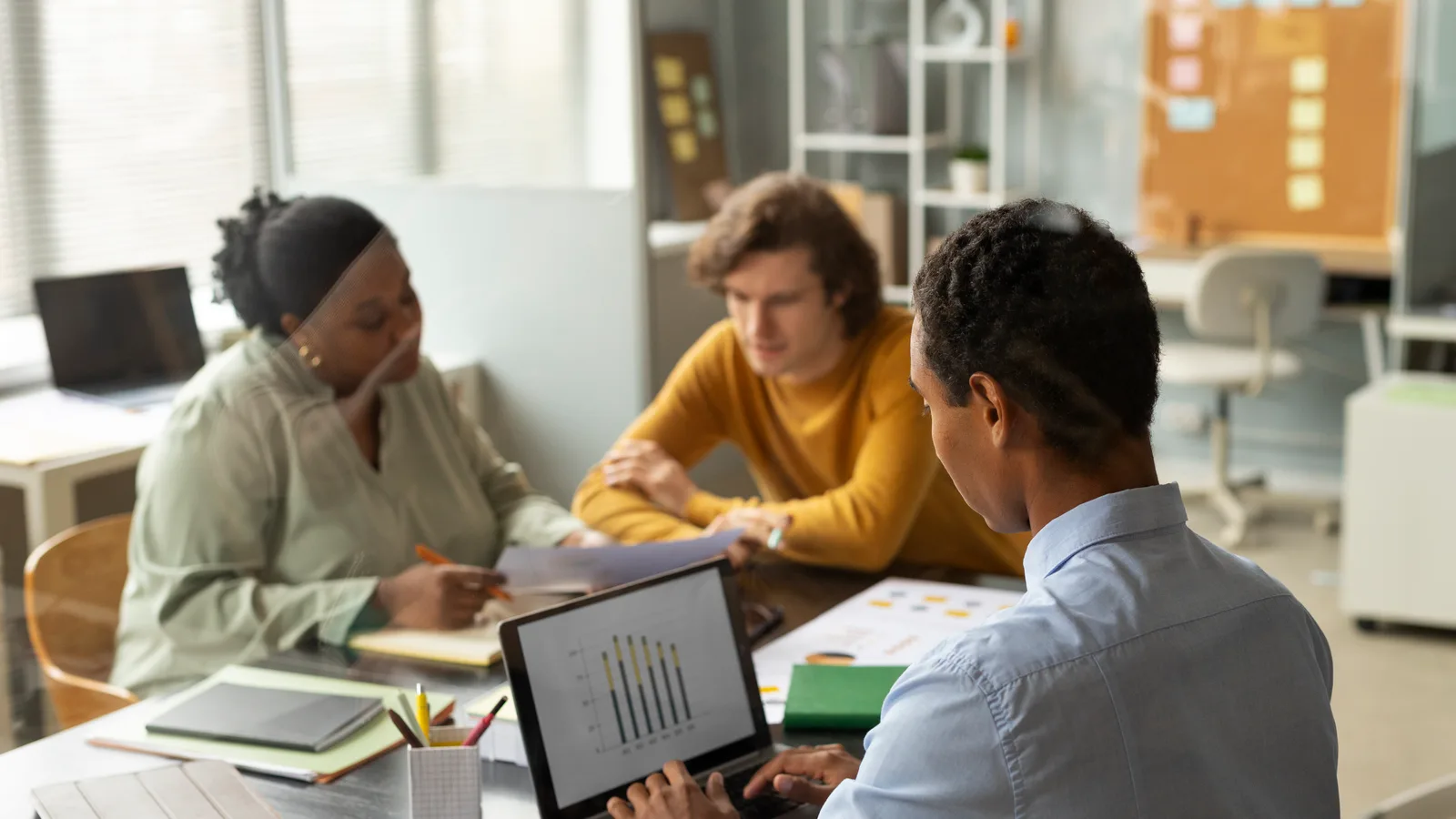 Three colleagues at a shared office table reviewing a bar chart on a laptop and printed notes