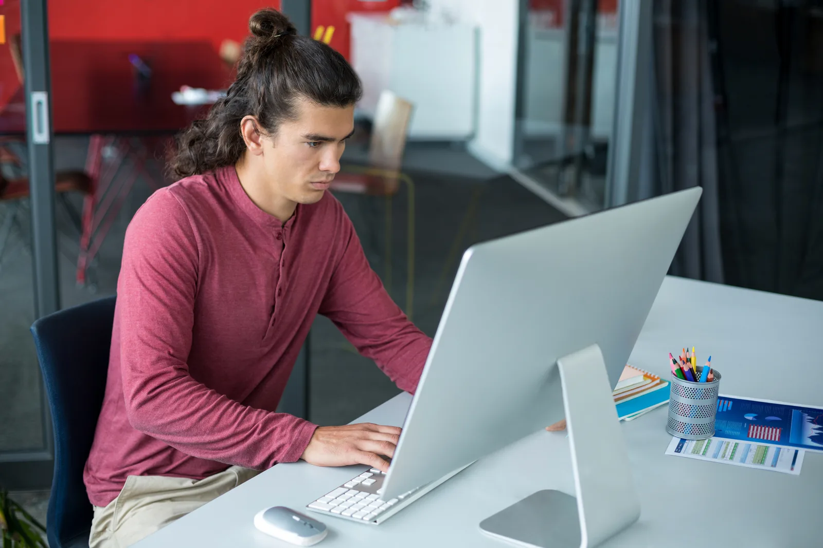 Young man at a desktop computer building a campaign, with printed reports and a pencil cup beside him