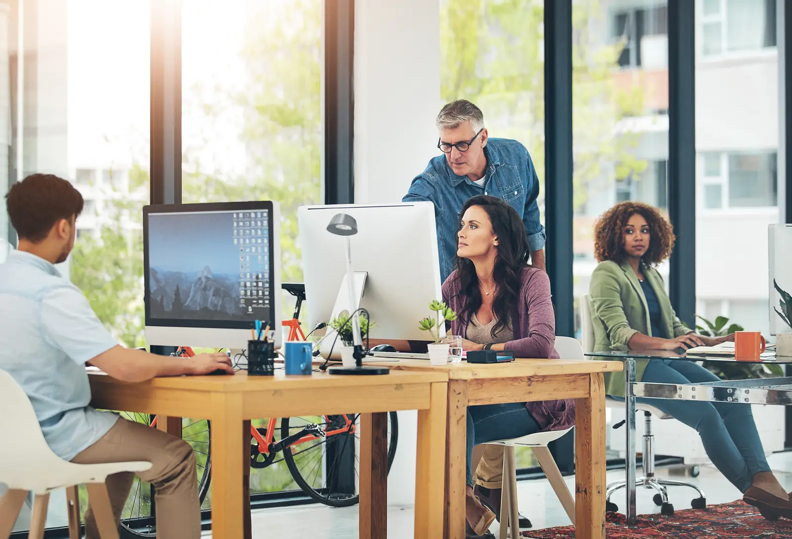A mentor leans over two colleagues at a desk, pointing at a client's screen in an open office