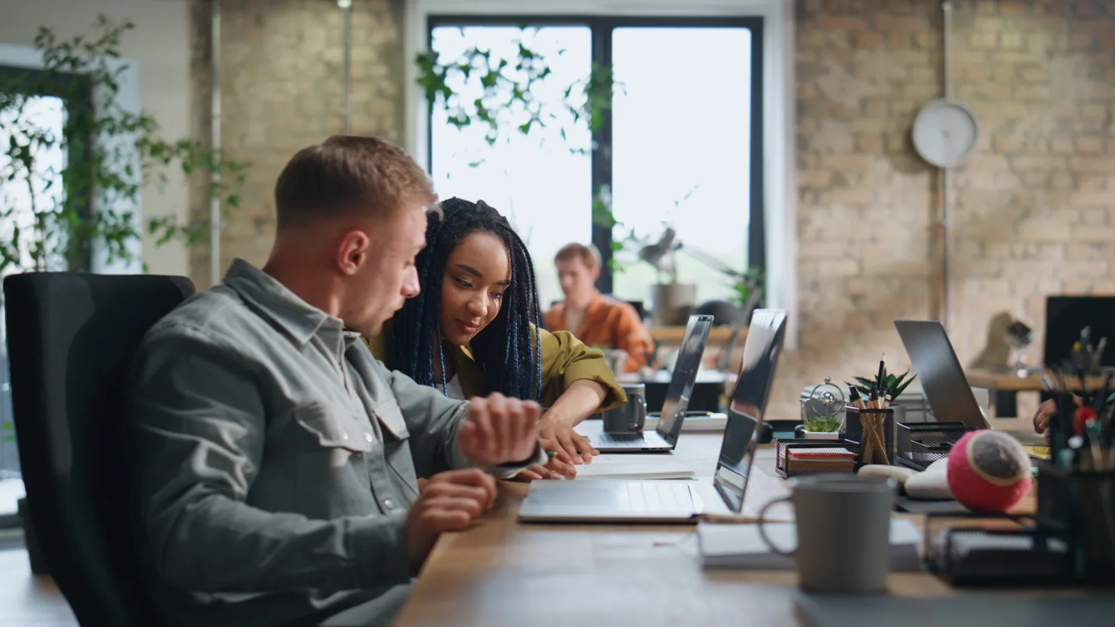 A woman at her desk asks a colleague about the account on her screen in an open office
