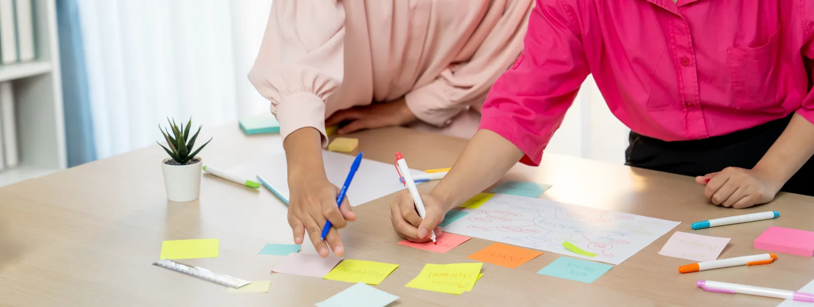 Two coworkers placing colored sticky notes onto a printed monthly calendar on a table