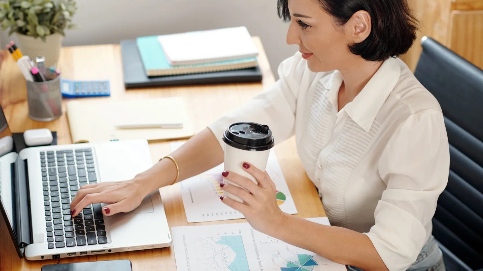 A woman types on her laptop at an office desk, coffee cup in hand, with printed charts and notebooks beside her