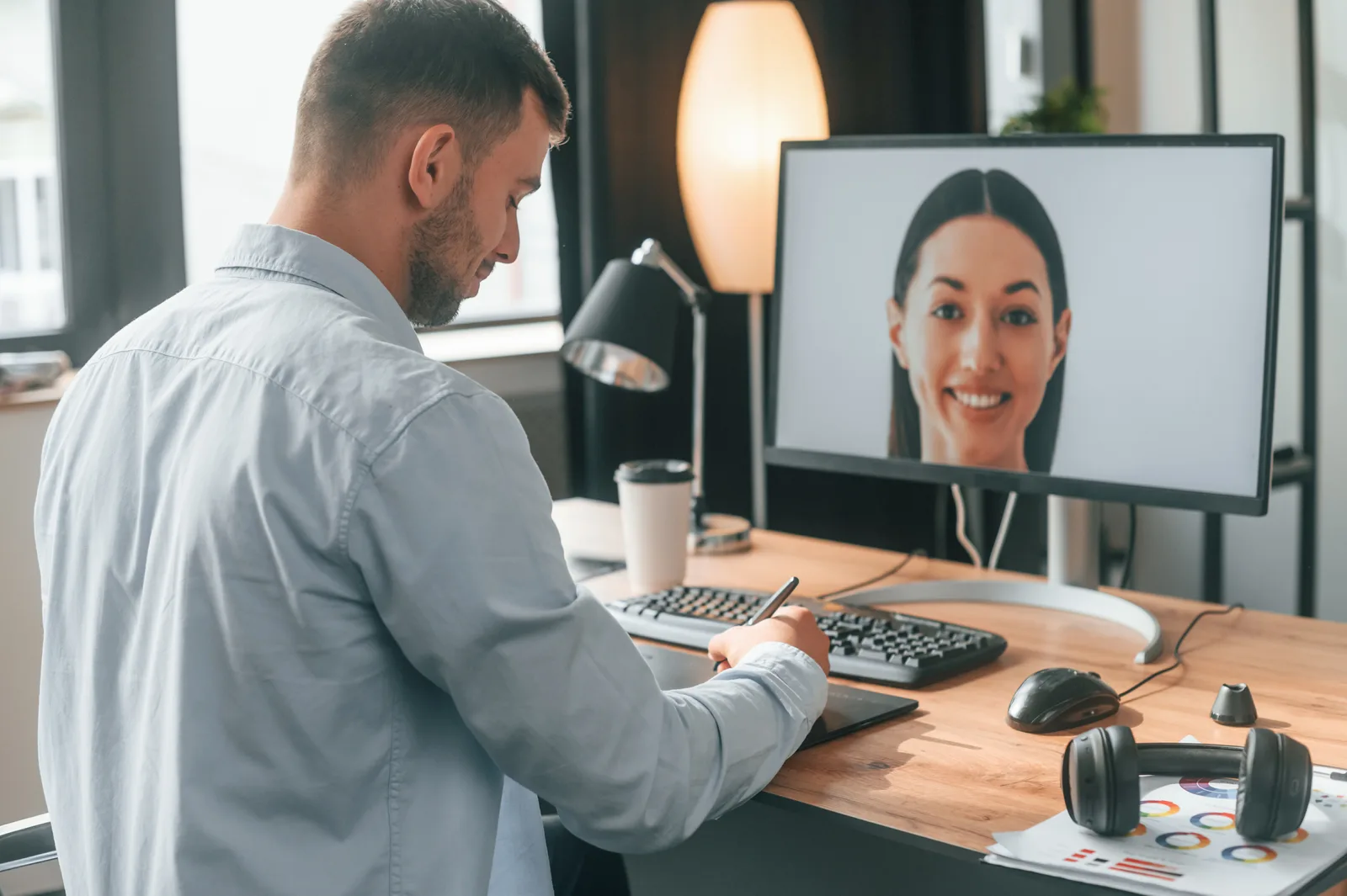 A man takes notes at his desk during a video call with a smiling woman on his monitor