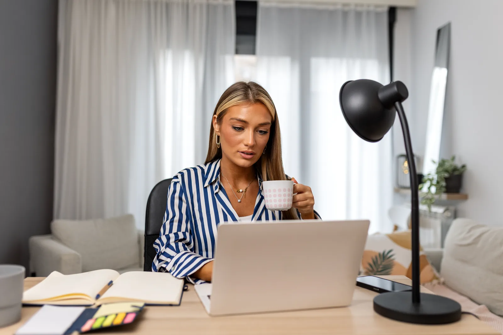 A woman sips from a mug as she works on her laptop at a home desk with an open notebook and sticky notes