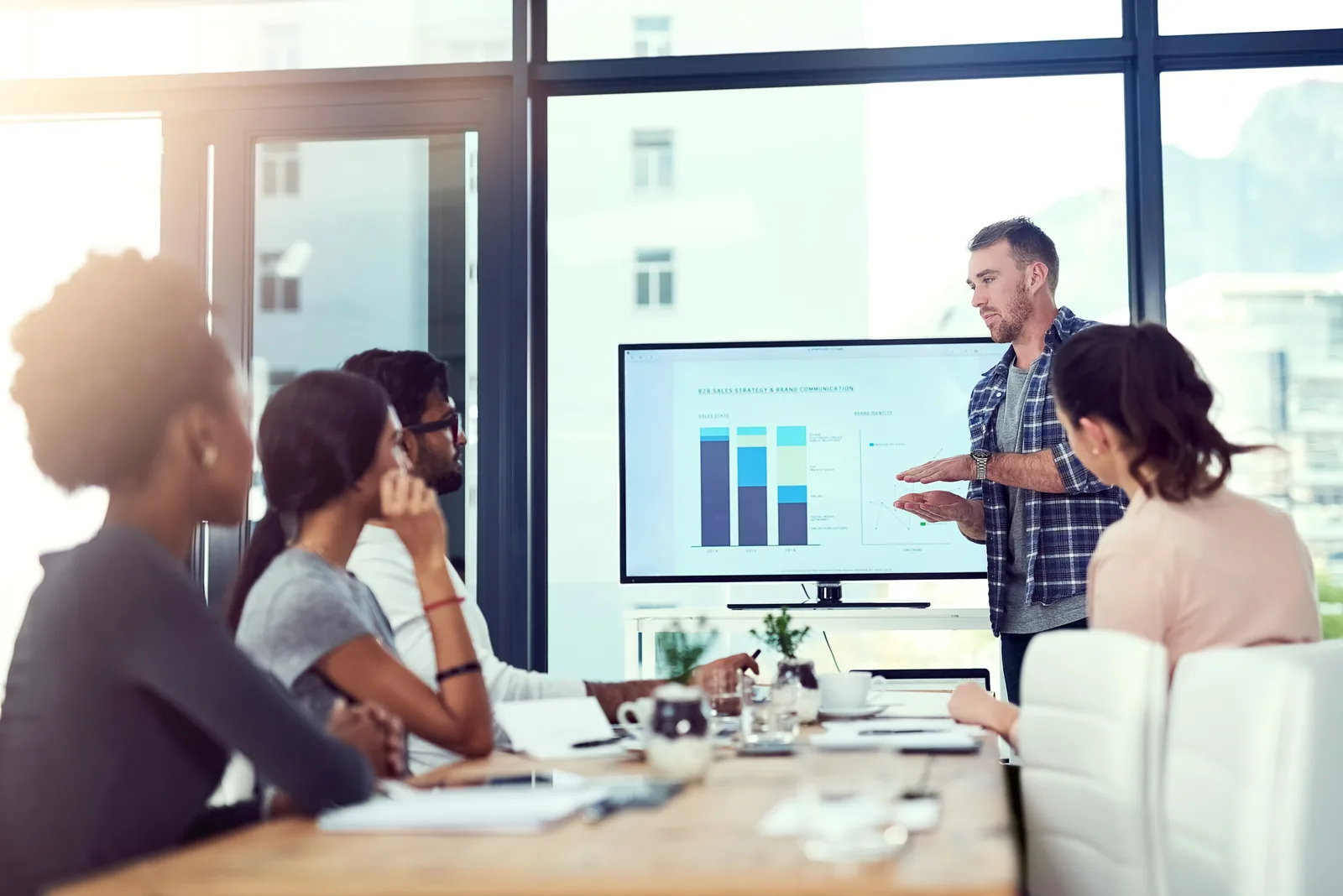 A manager walks four colleagues through a bar chart on a wall screen in a bright glass-walled meeting room