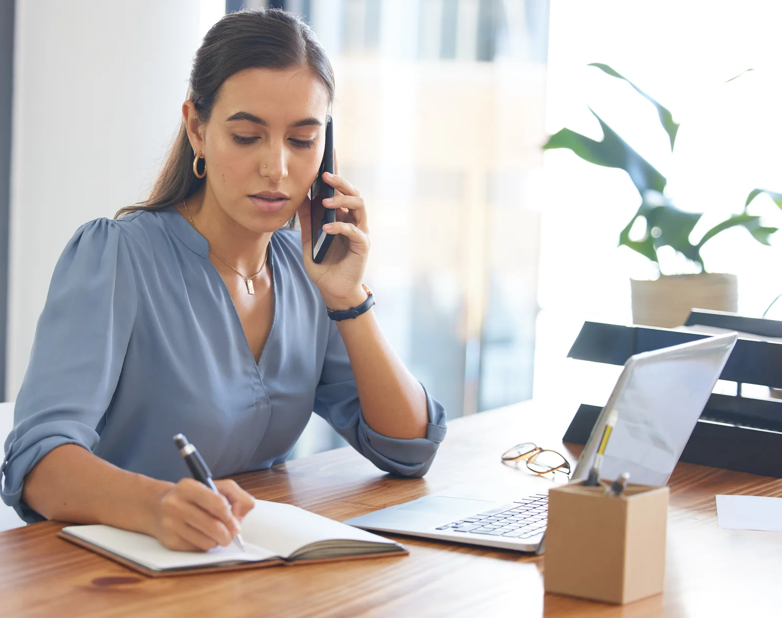 An executive assistant on a cell phone call writes in an open notebook beside her laptop, glasses set down on the desk