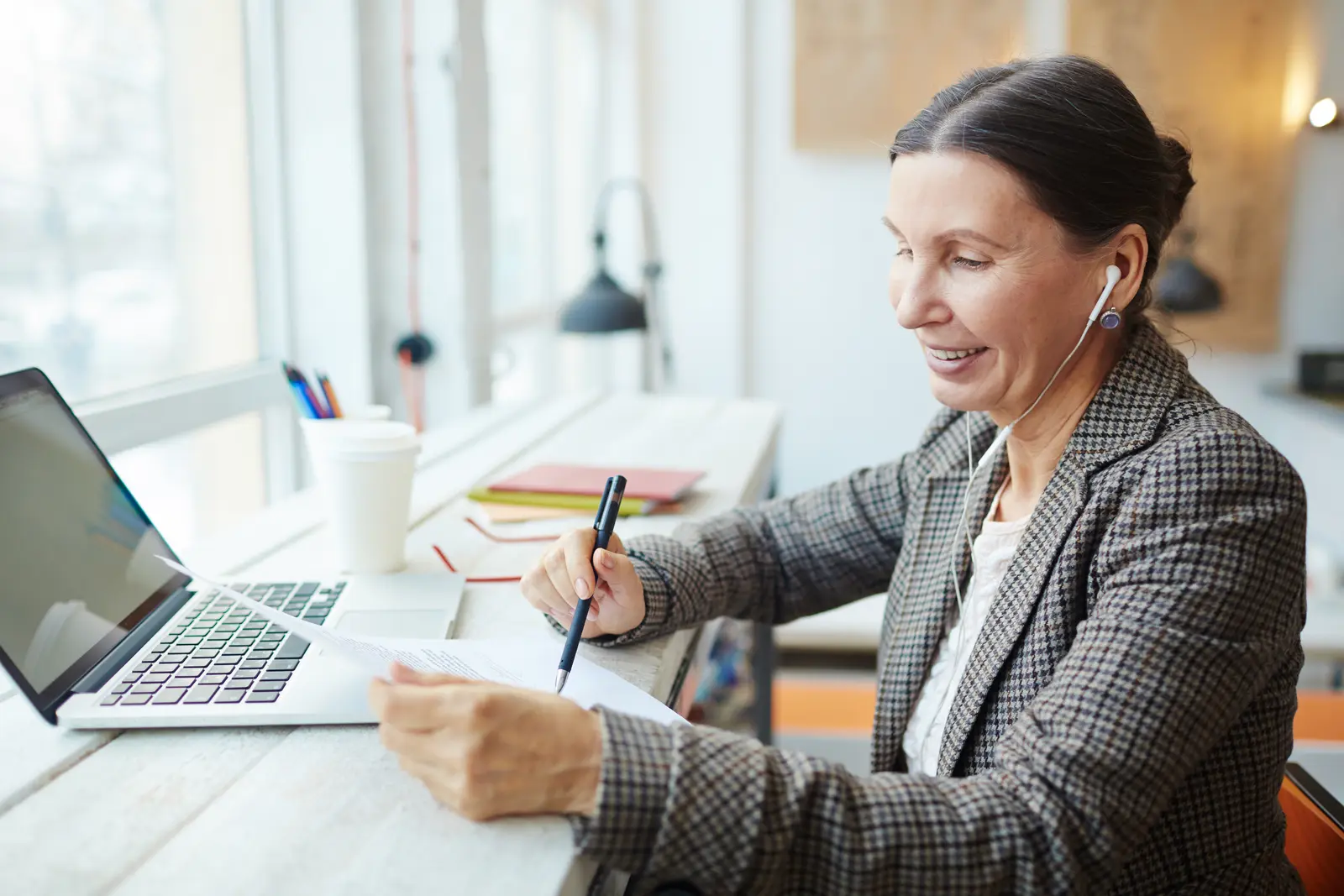 A business owner listens through earbuds and marks up printed pages beside her open laptop at a window desk