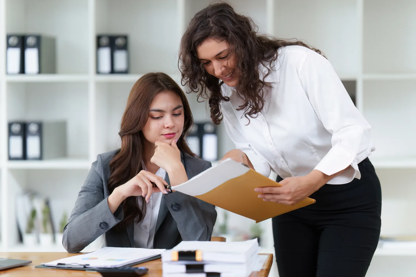 A founder leans in to hand a manila folder of papers to her new assistant, who sits at a desk stacked with files