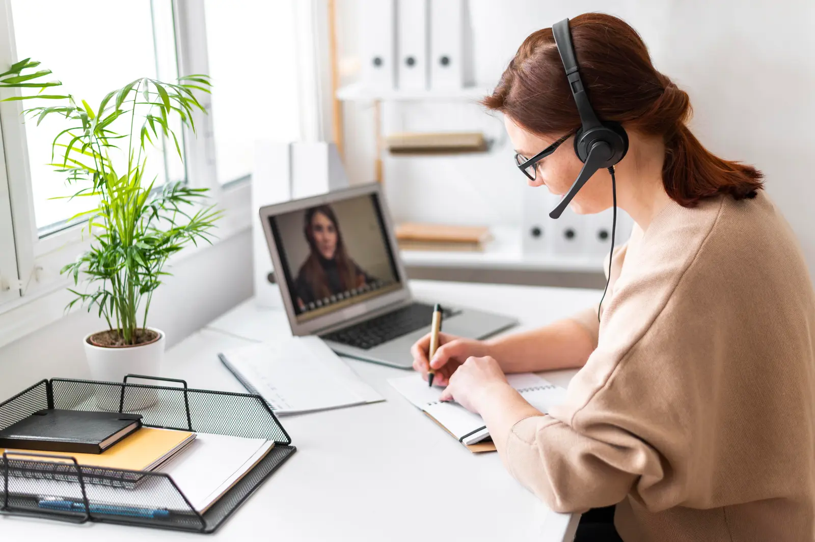 A remote executive assistant in a headset takes notes during a laptop video call at a white desk by a sunny window