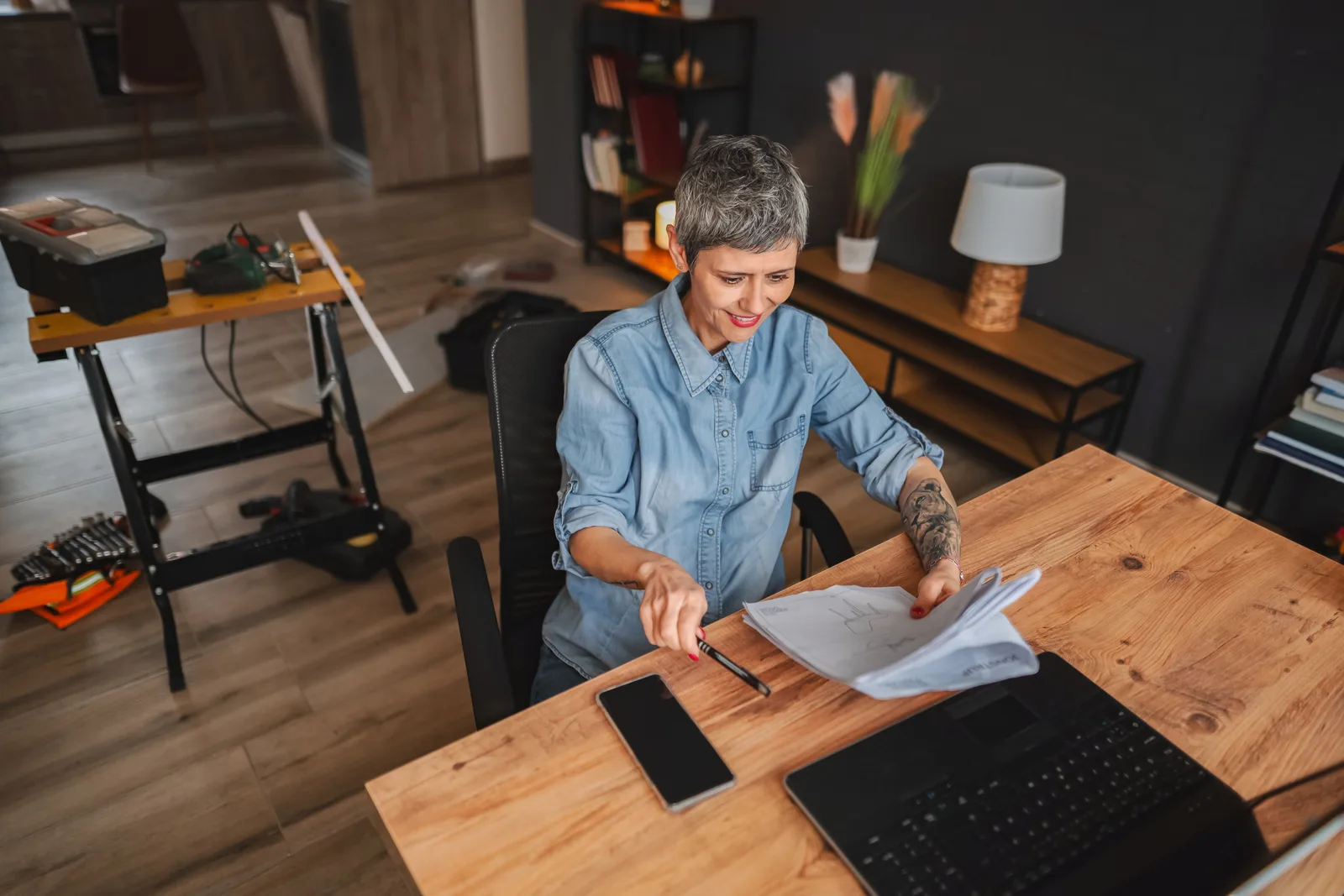 A business owner compares printed plans at her home office desk, laptop open beside them