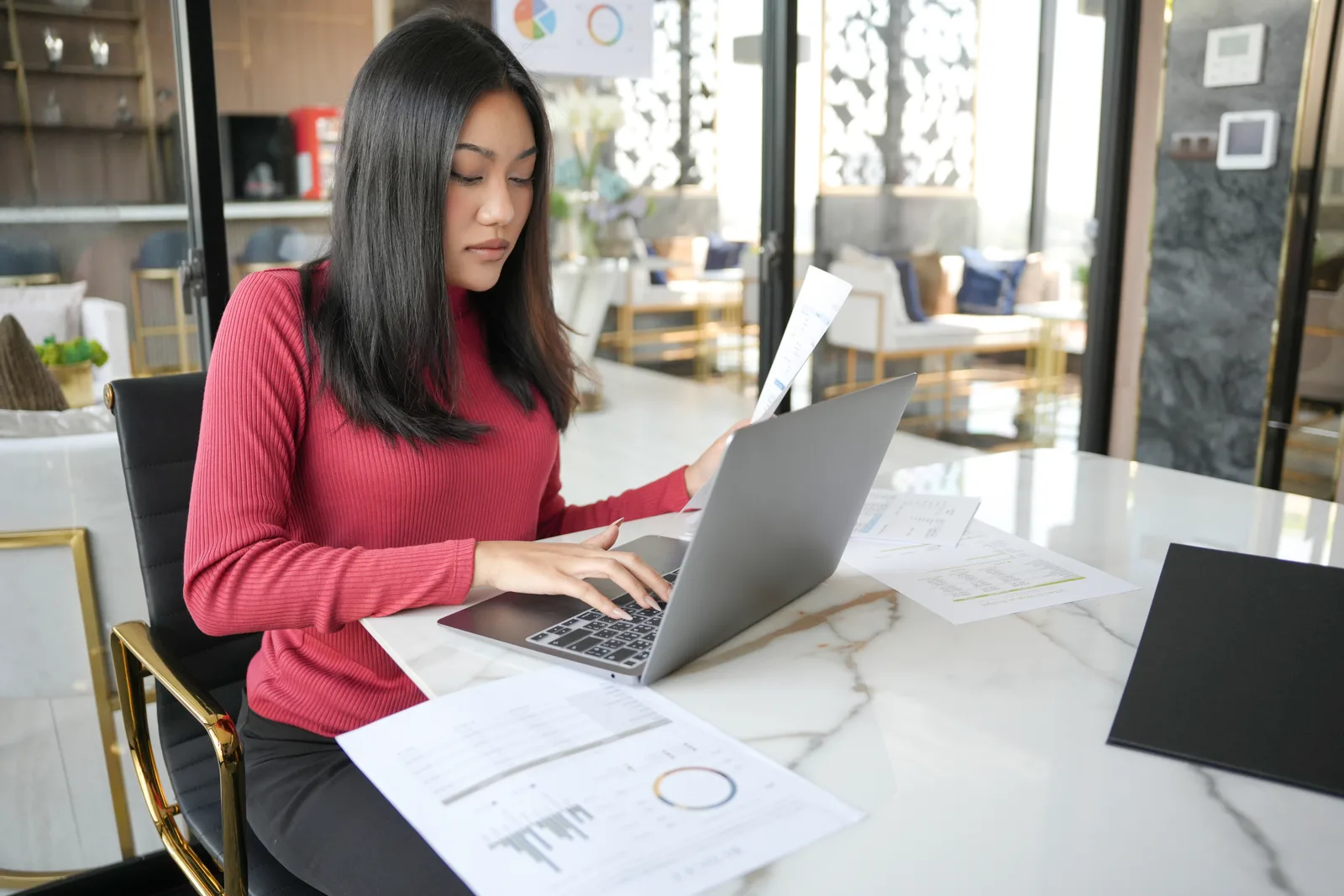 A woman at a laptop in a bright office checks printed charts against the screen before deciding