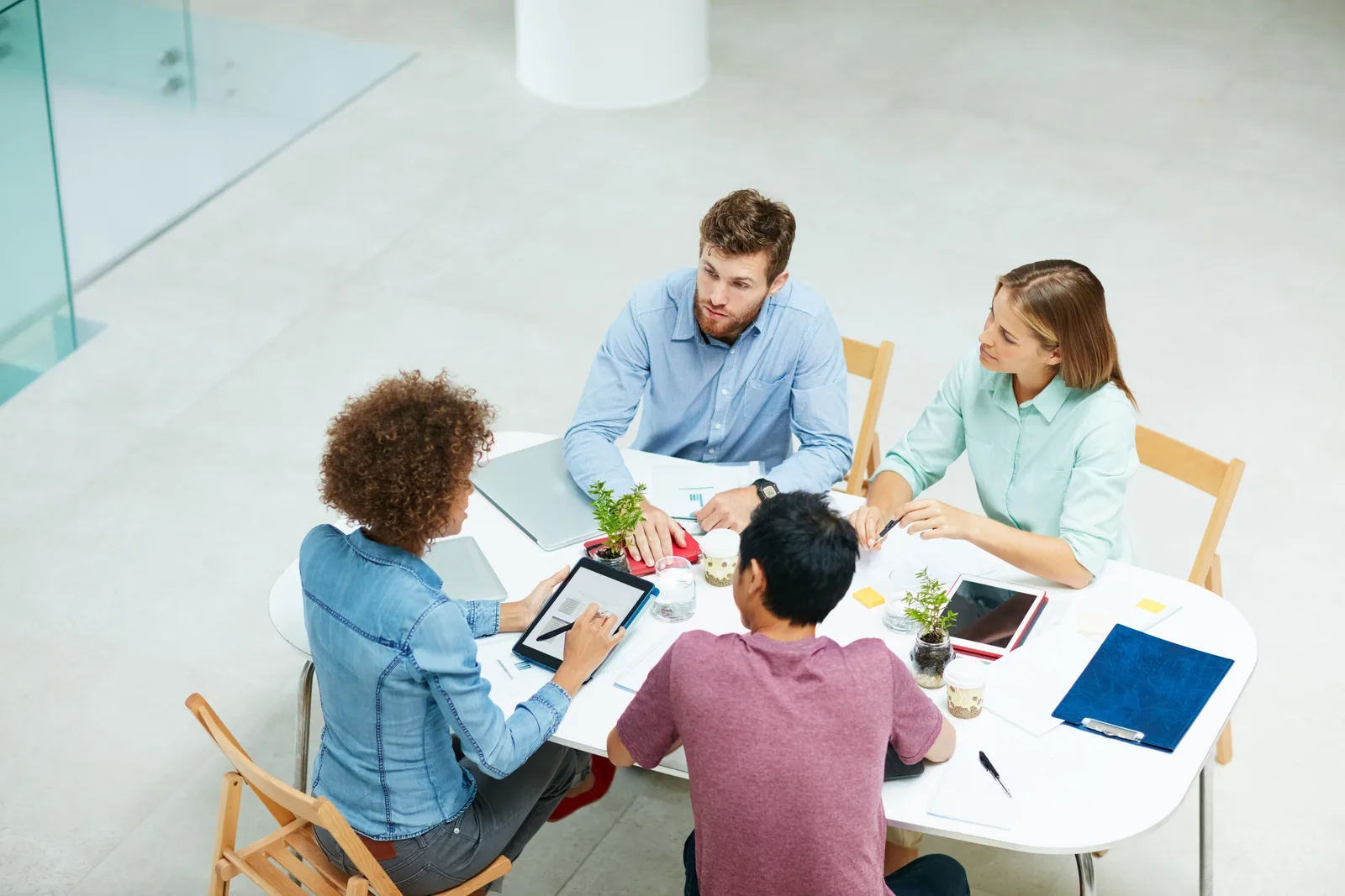 Four colleagues around a white table talk through options on laptops and paper in a bright office