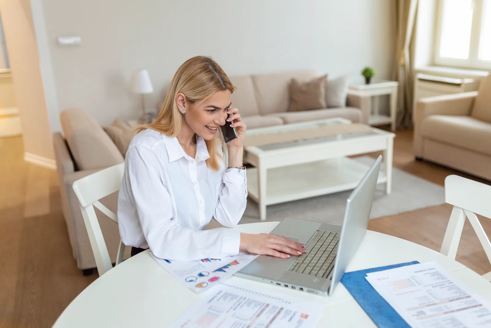 A real estate agent on a call at her laptop in a staged living room, papers spread across the table