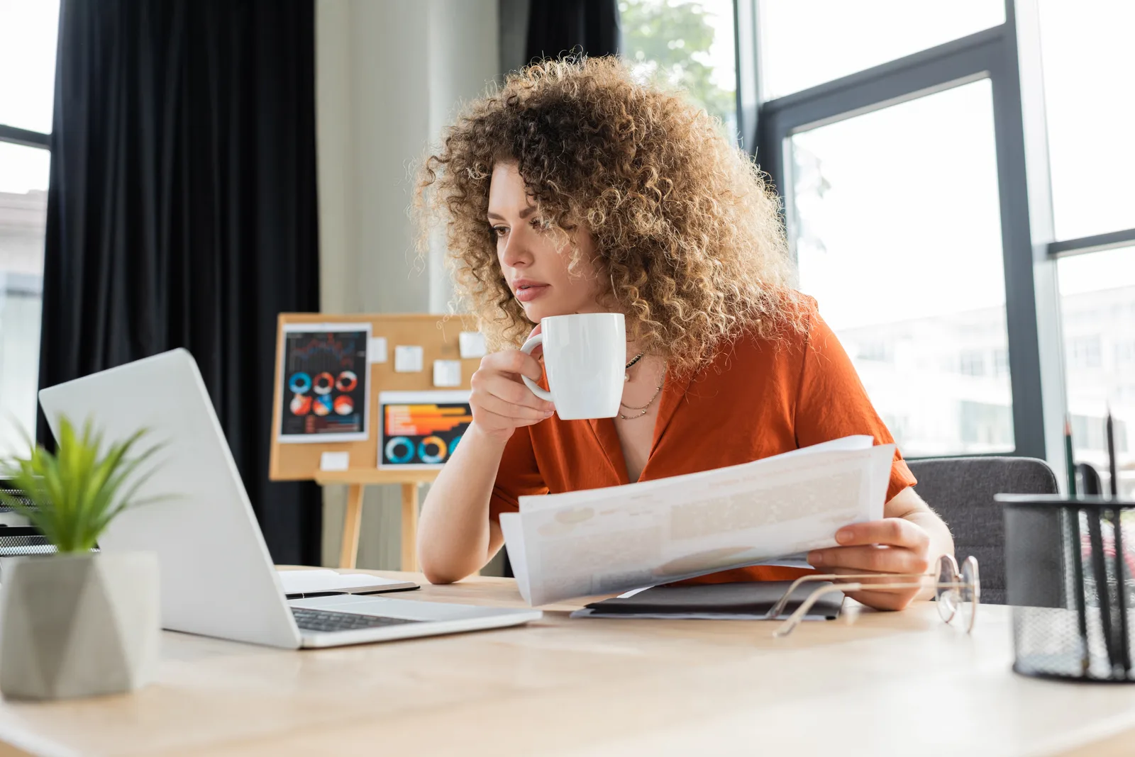 A businesswoman with a cup of tea reads a printed report at her laptop