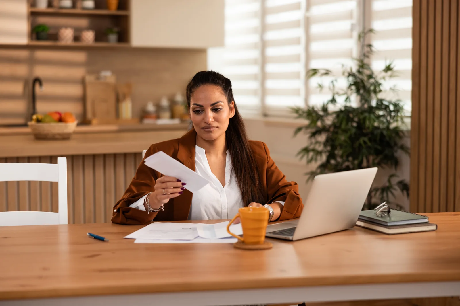 A businesswoman at her kitchen table reads a printed statement beside her laptop