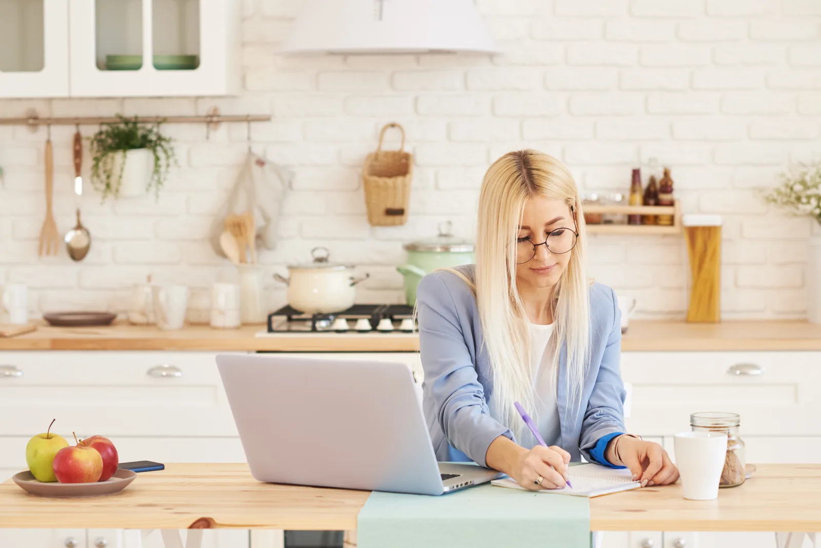 A woman in glasses takes notes beside her open laptop at a bright kitchen table