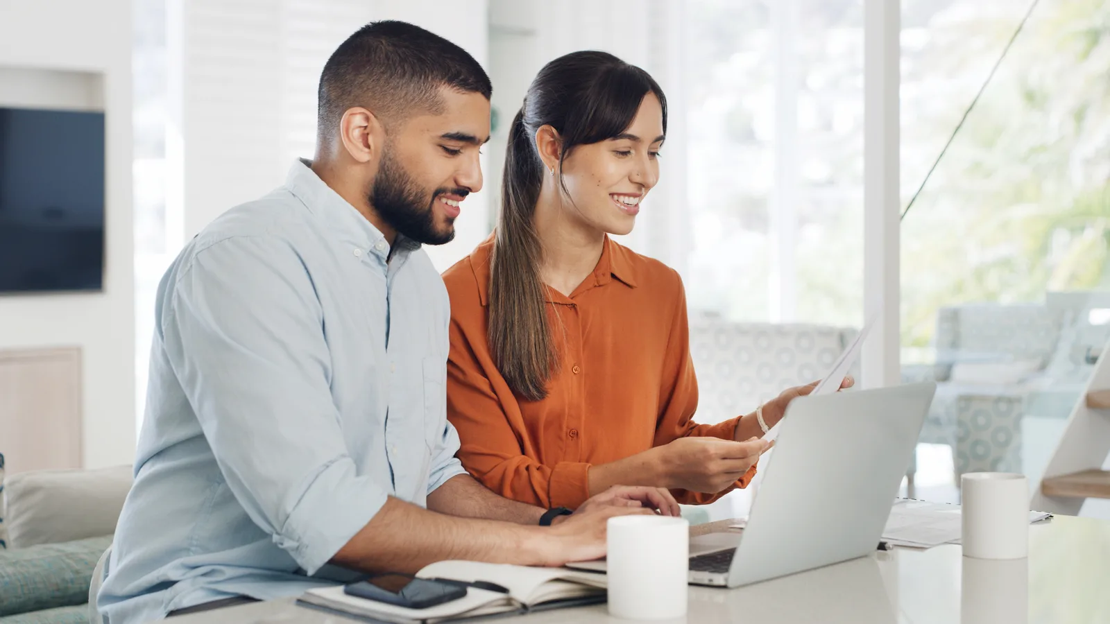 Two business partners compare a printed quote against a laptop at a bright kitchen table
