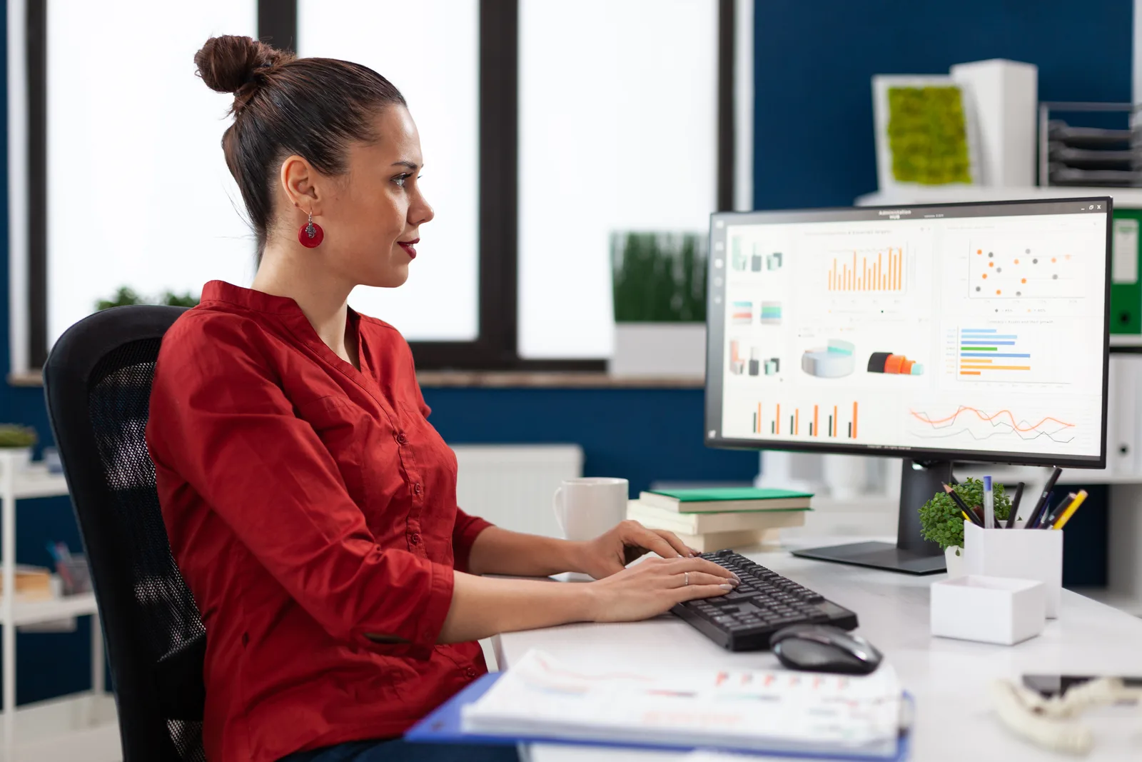 A manager in a red shirt studies a monitor full of bar charts and trend lines at her office desk