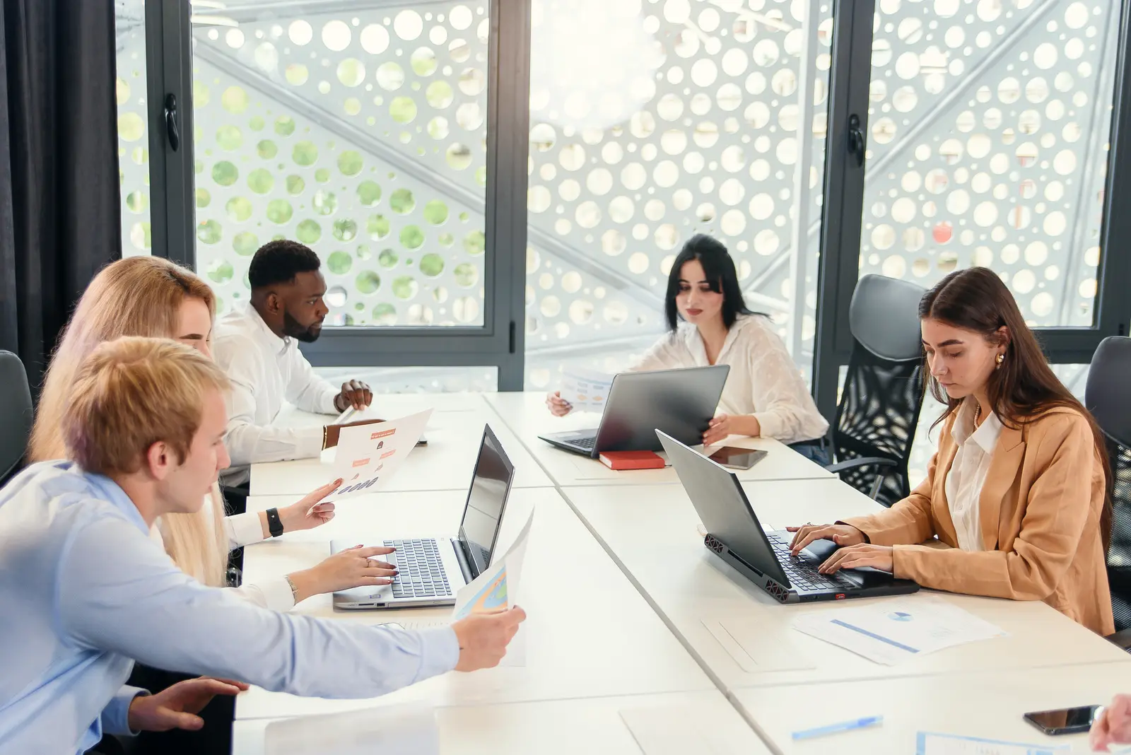 Five colleagues work at laptops around a long white table in a bright glass-walled meeting room