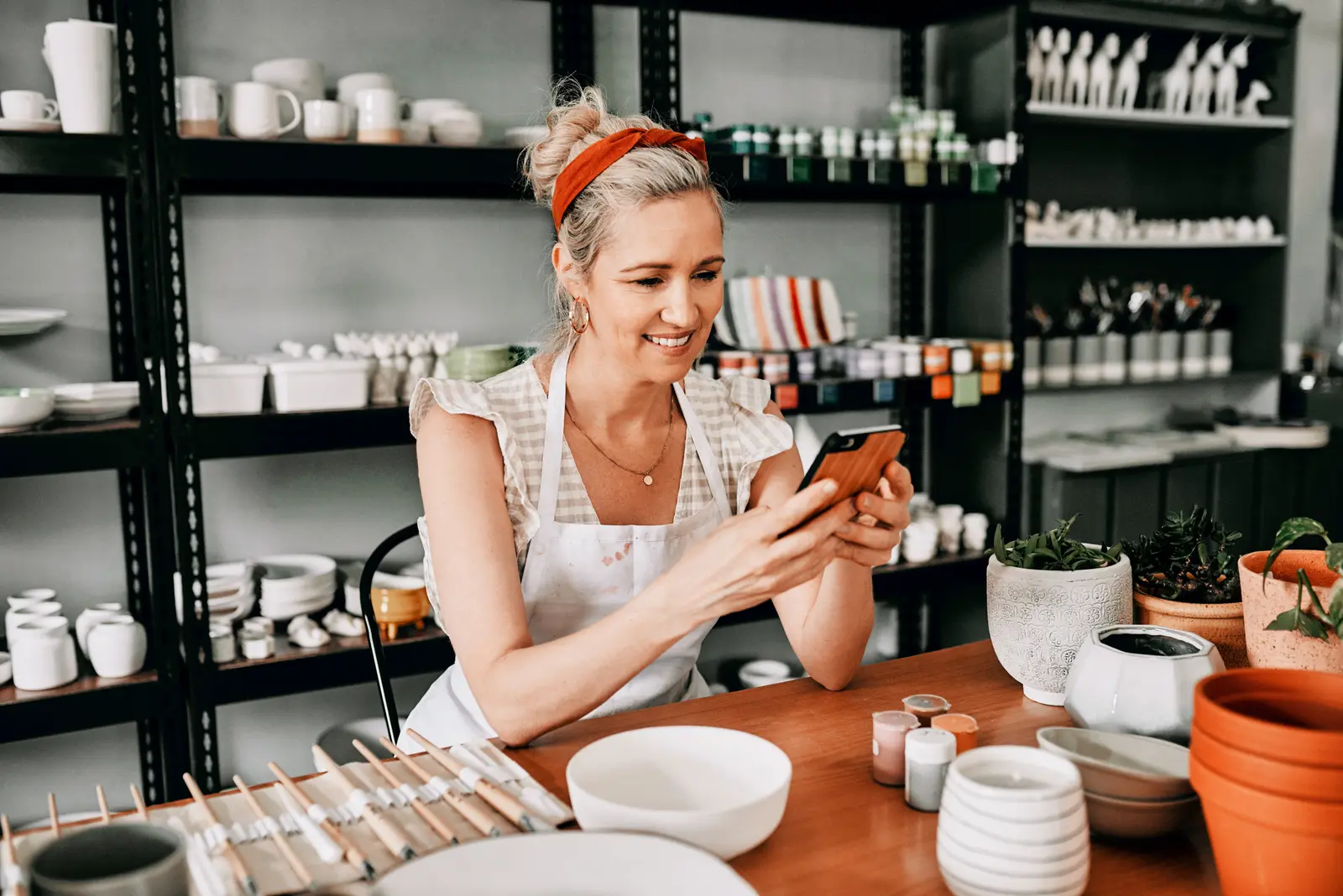 A pottery studio owner in an apron smiles at her phone at a workbench stacked with bowls and glazes