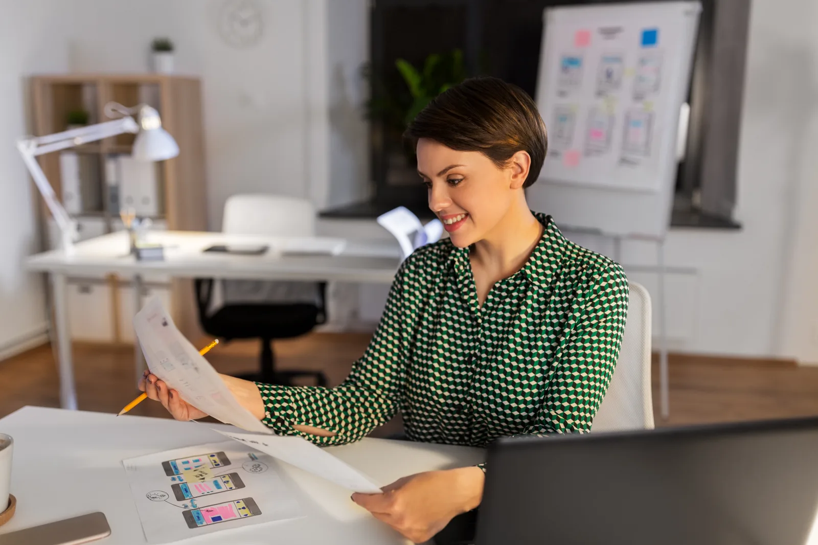 A designer smiles at printed app layouts at her desk, pencil in hand, more layout sketches beside her laptop
