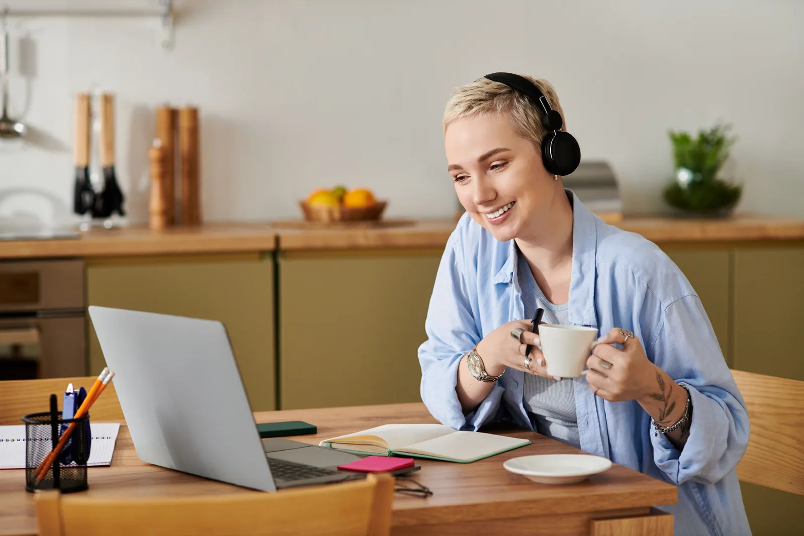 A remote worker in headphones smiles at her laptop, coffee cup in hand and a notebook open on the kitchen table