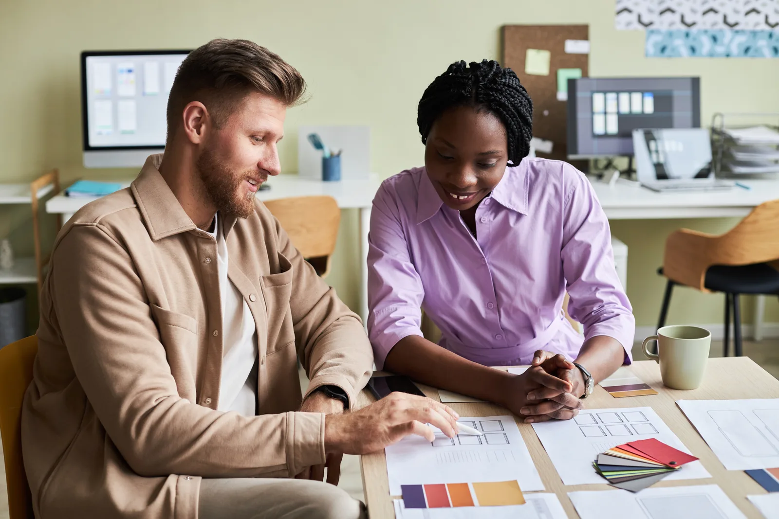Two colleagues go over printed page layouts and color swatches spread across a shared table