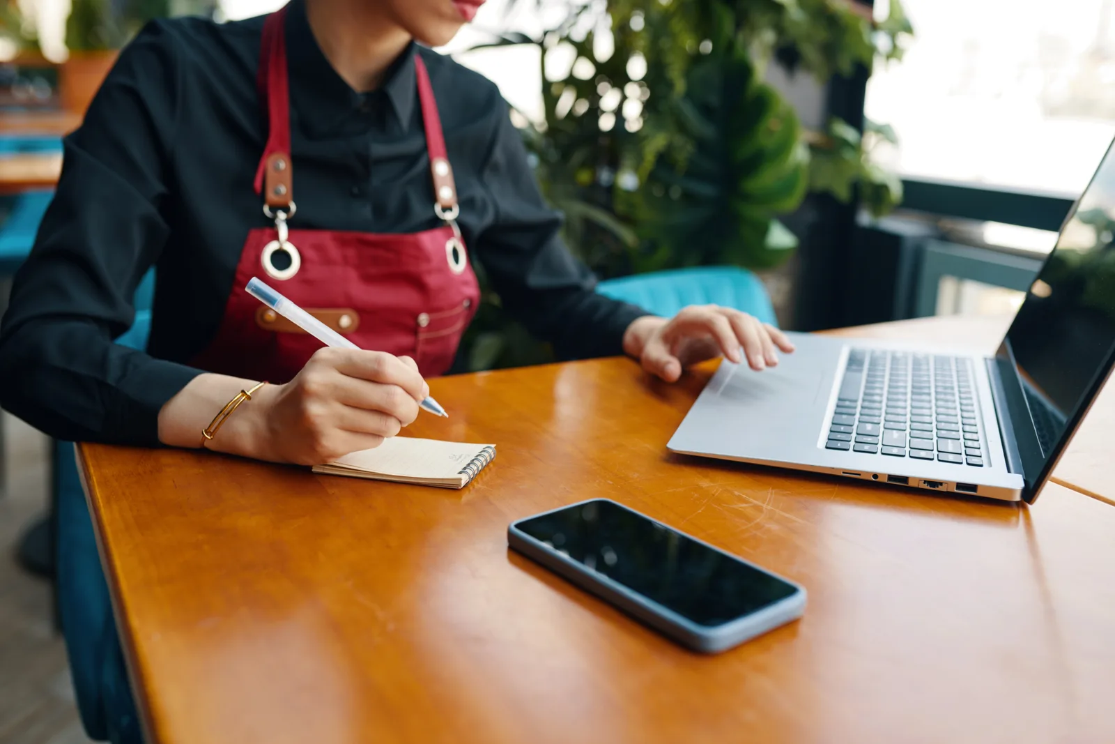 A woman in a red apron writes in a small notepad beside her laptop and phone at a cafe table