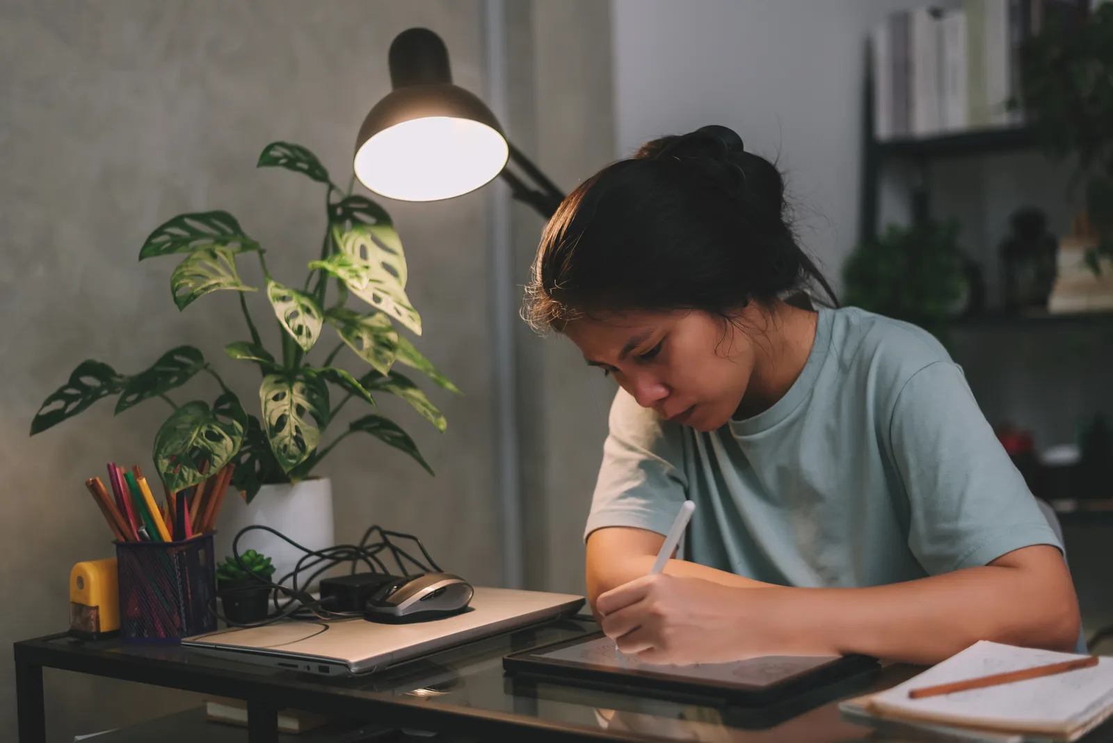 A freelance graphic designer sketches on a tablet with a stylus under a desk lamp, working late at home
