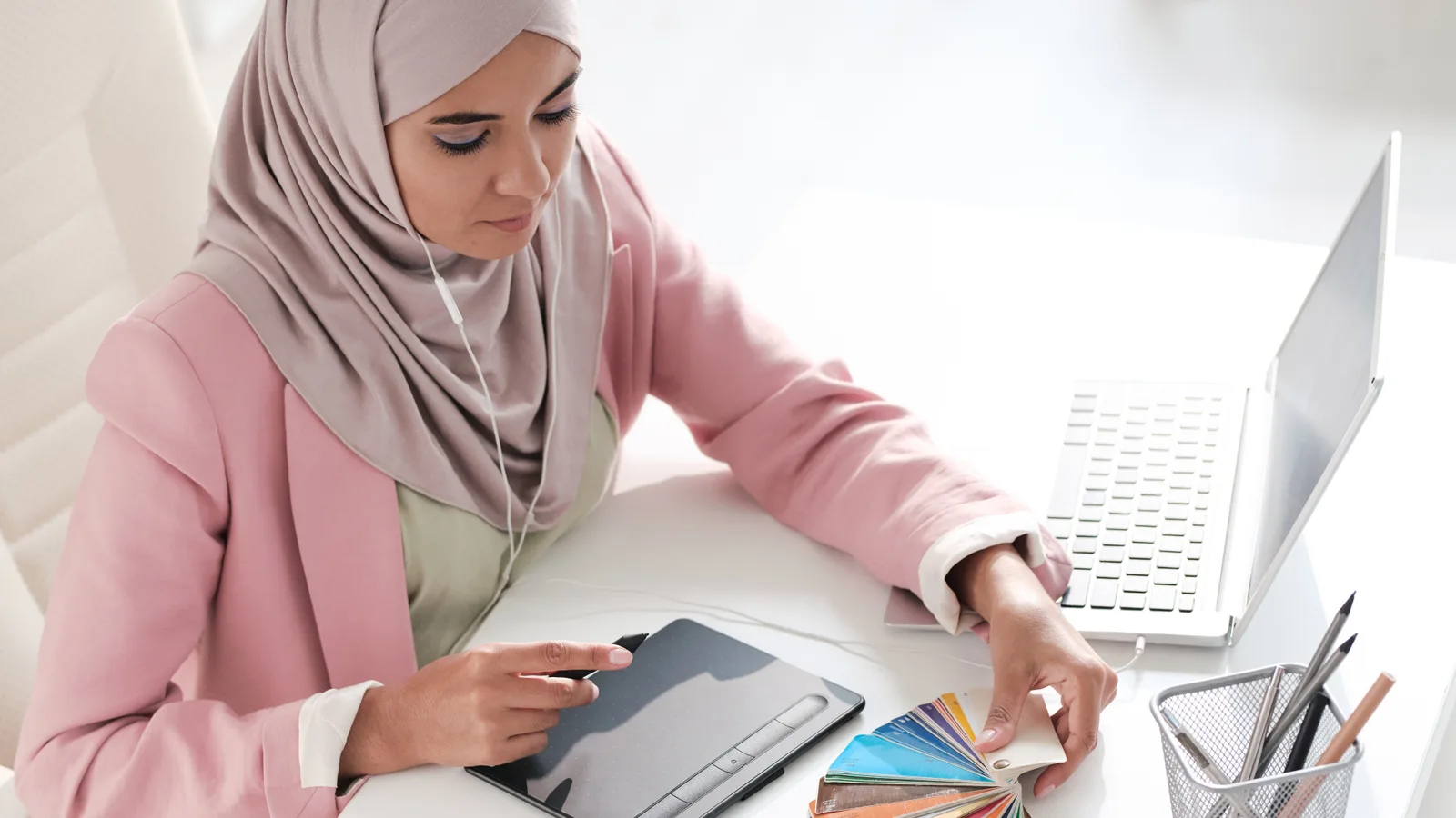 A graphic designer picks colors from a swatch fan beside her pen tablet and laptop in a bright white office