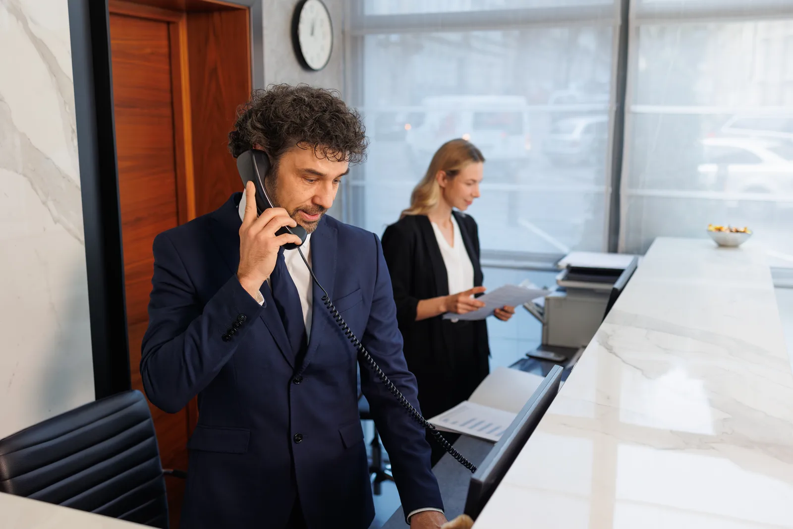 A hotel receptionist takes a call on a landline at the front desk while a colleague works behind him