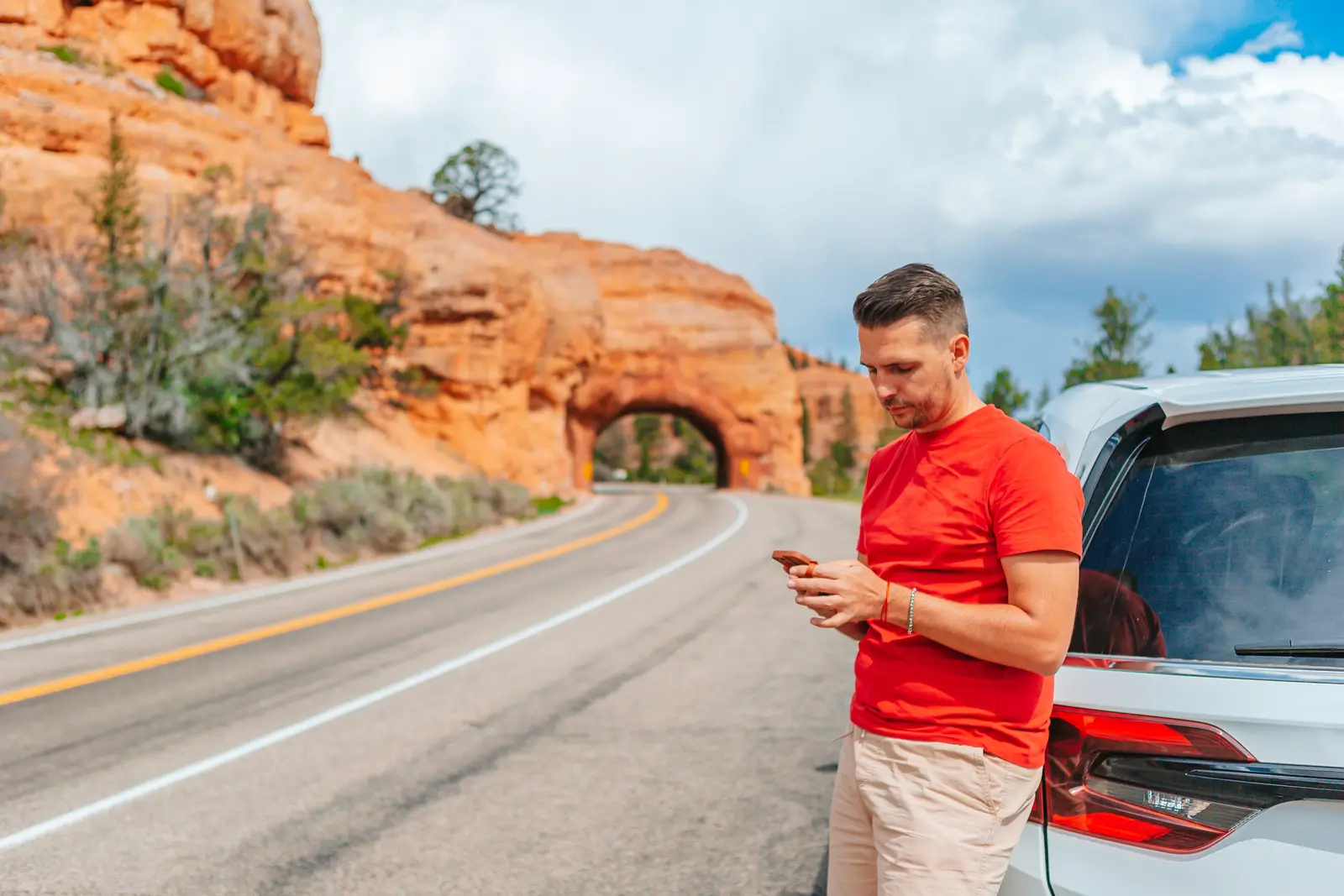 A traveller stands beside a parked car on a desert canyon road, checking his phone