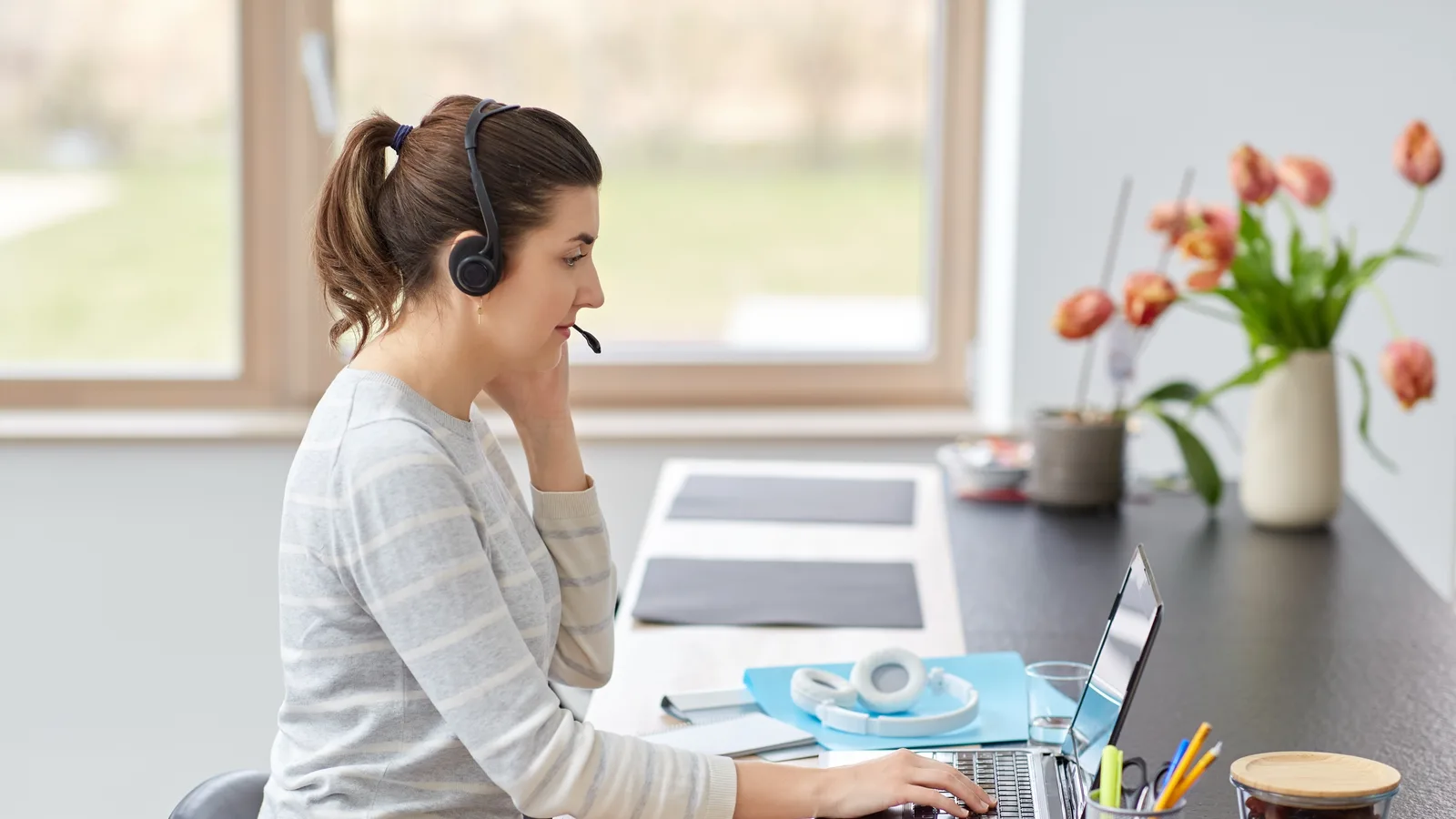 A virtual assistant in a headset types on her laptop at a bright home desk by the window