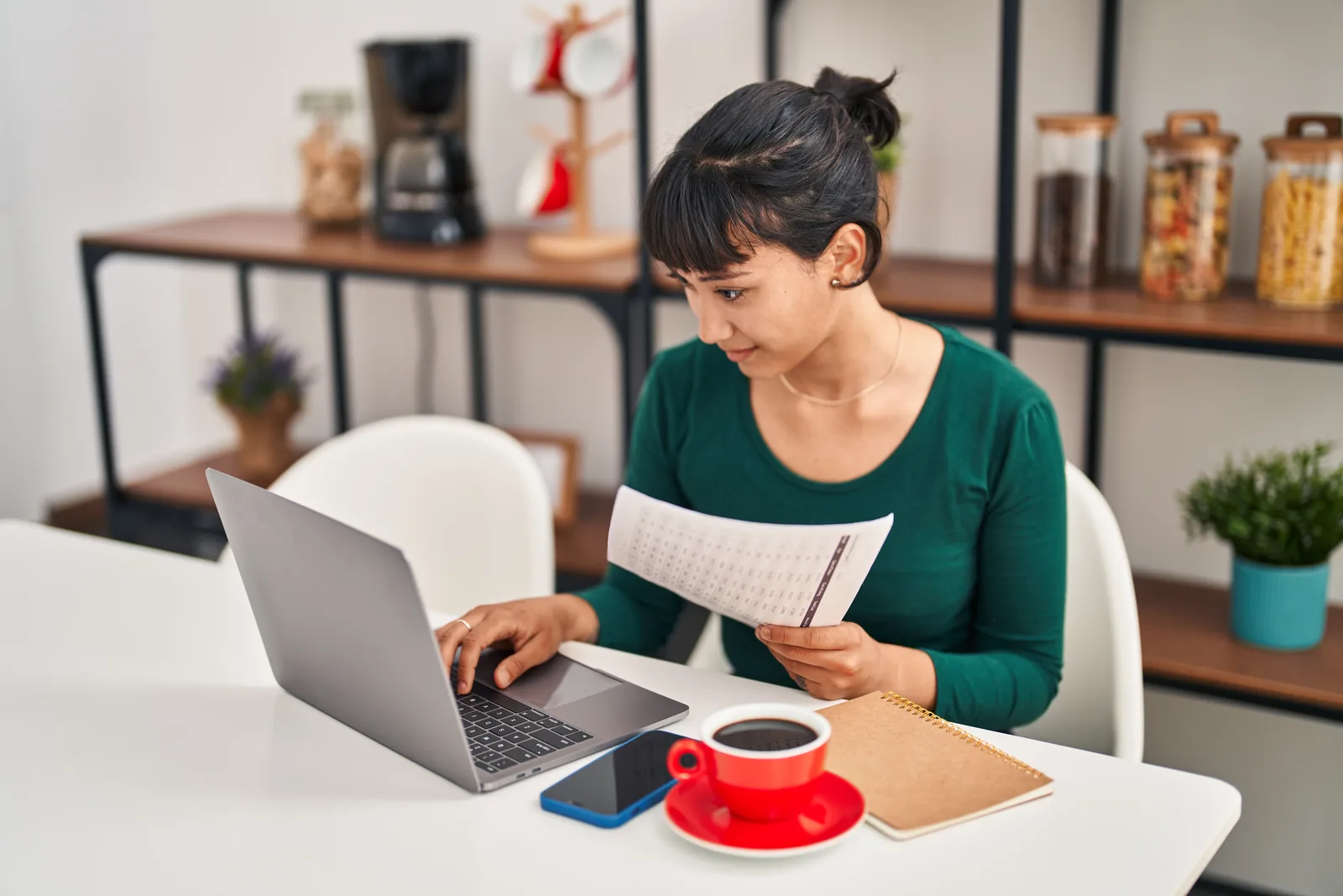 An assistant holds a printed statement and checks it against her laptop screen at a home table