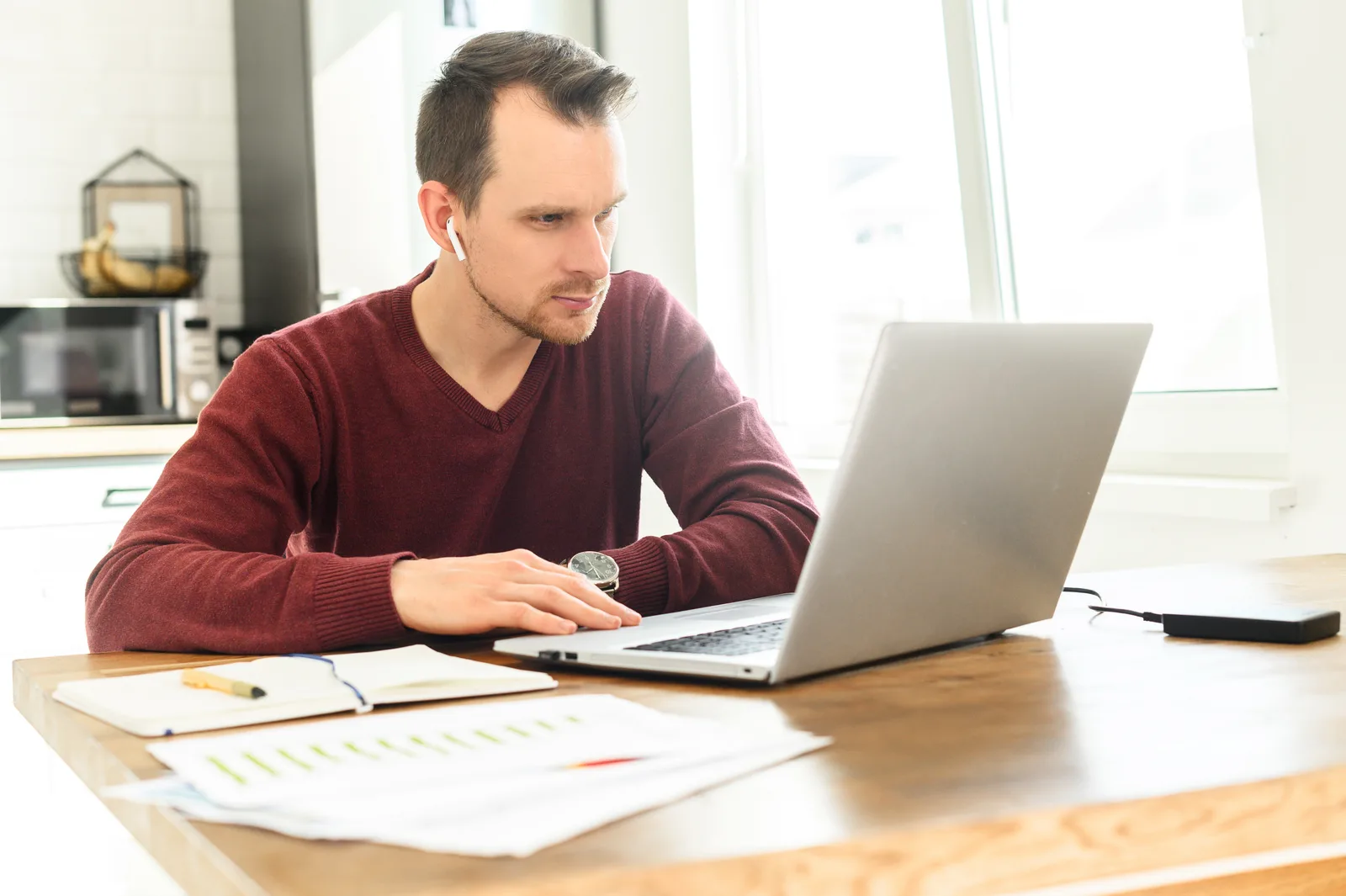 A man wearing an earbud works on his laptop at a kitchen table, a notebook and printed charts beside him