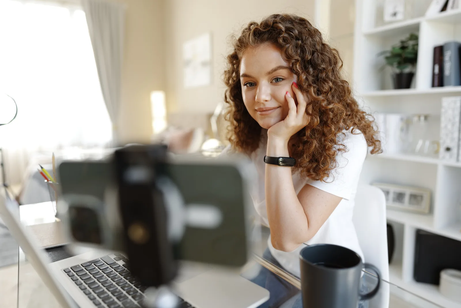 A woman records herself on a phone camera mounted at her home desk, her laptop open in front of her