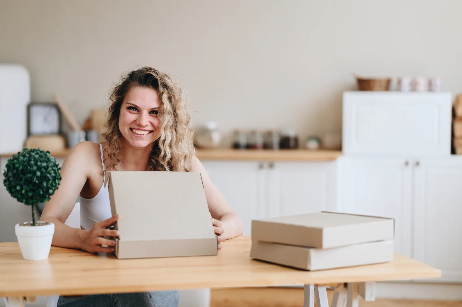 A smiling woman opens a flat cardboard box at a wooden table at home, two more boxes stacked beside her