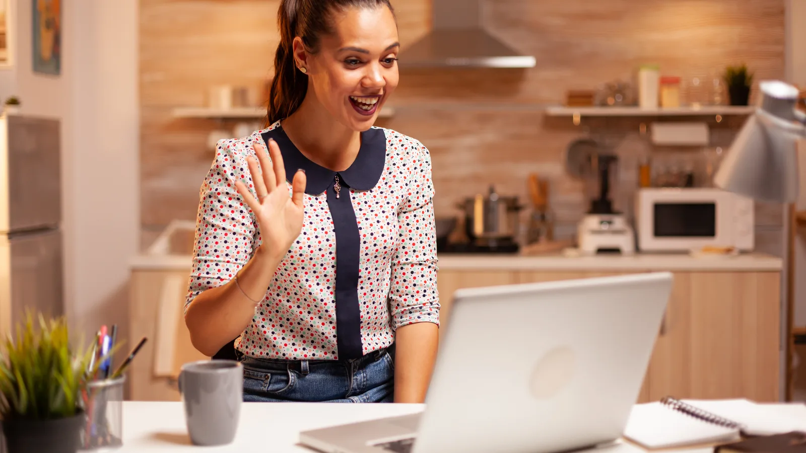 A woman working from home waves at her laptop on a video call from her kitchen table, a mug and notebook beside her